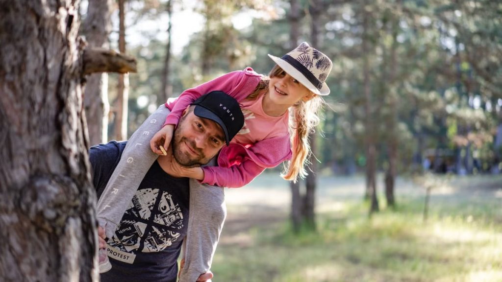 A joyful father giving his daughter a piggyback ride in a sunny forest setting.