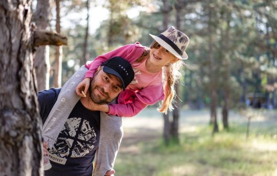 A joyful father giving his daughter a piggyback ride in a sunny forest setting.